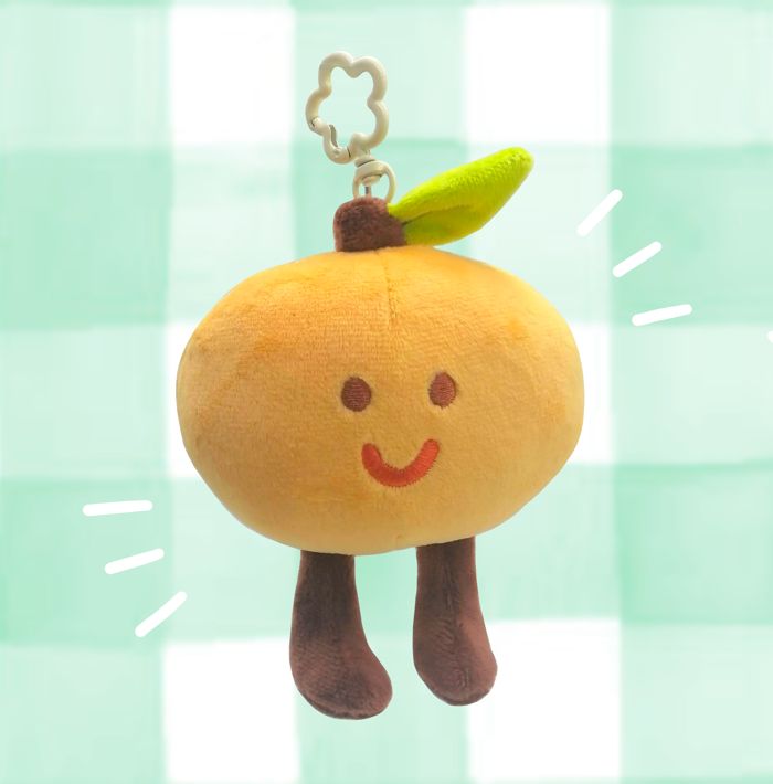 a plush of a little orange keychain with a white flower hook. it has brown feet dangling and a happy little smile. 