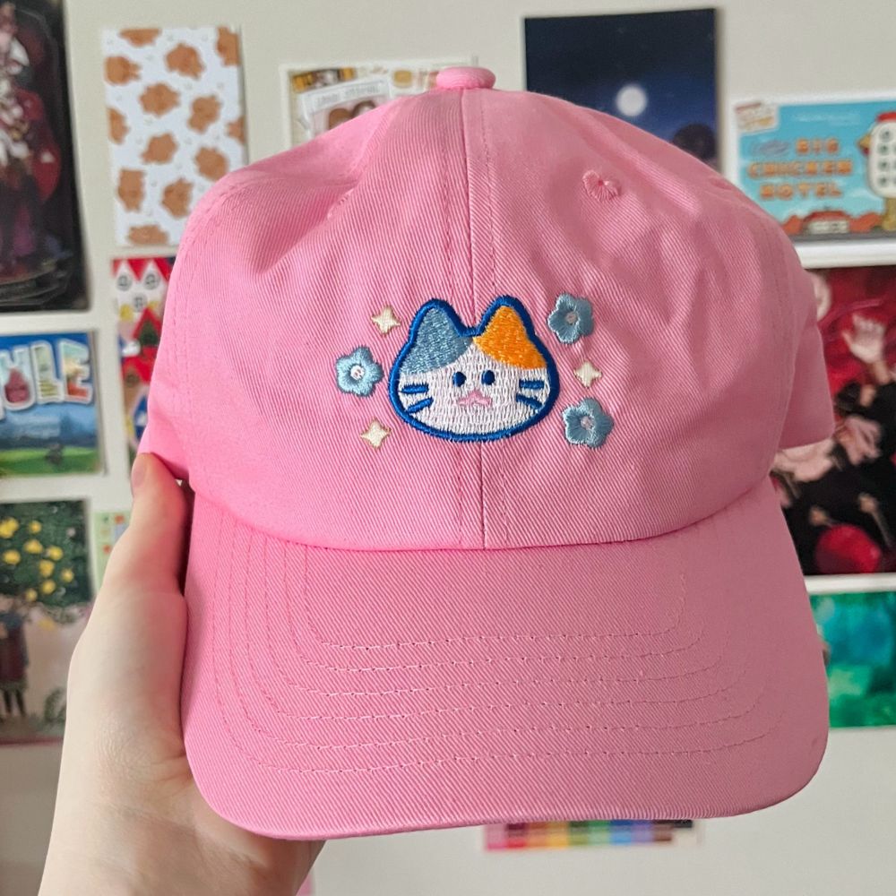 a pink cap with an embroidered pastel calico face on it, surrounded by blue flowers and light gold sparkles. 