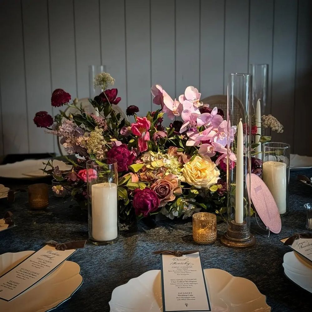 An elegant floral centerpiece on a dark linen table with blush orchids, deep magenta ranunculus, golden yellow roses, and soft greenery, surrounded by taper candles and scalloped white plates.