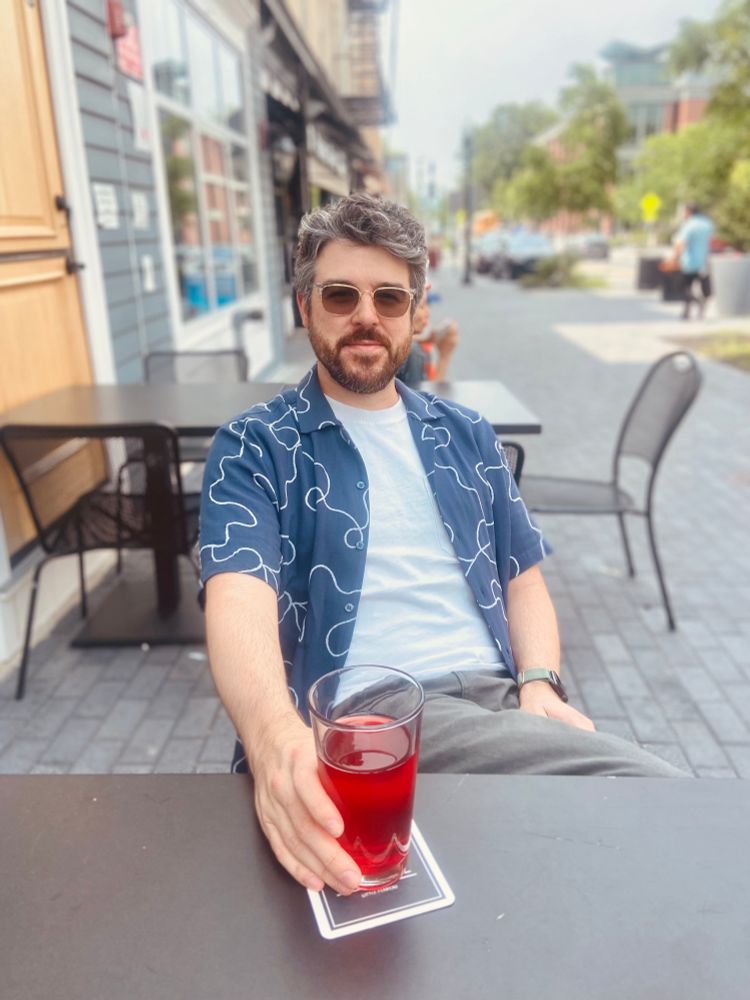Me sitting outside along a street in a white t shirt with a blue t shirt over it having a red colored beer.