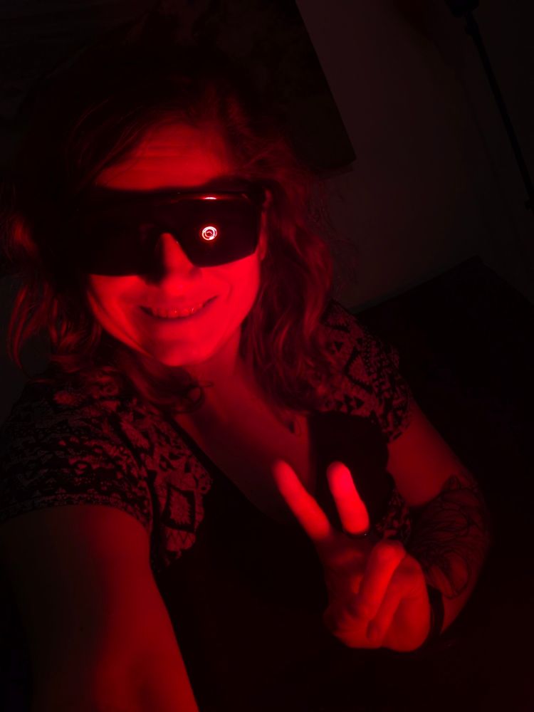 Ich sitze auf der Couch. Ich trage eine große schwarze Verdunkelungsbrille, um das Augenlicht gegen das Rotlicht zu schützen. Offene blonde Haare. Meine linke Hand zeigt ein Victory in die Kamera. Ich lächle in die Kamera. Auf dem linken Brillenglas sieht man das reflektierte Rotlicht. Das Bild ist sehr dunkel, in einem roten Ton, gehalten. 