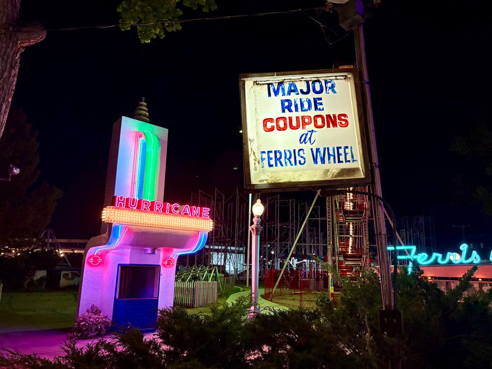 A very well worn lit sign saying “major ride coupons at Ferris wheel” that has seen better days, and more neon in the background - one for the Hurricane and one for the Ferris wheel