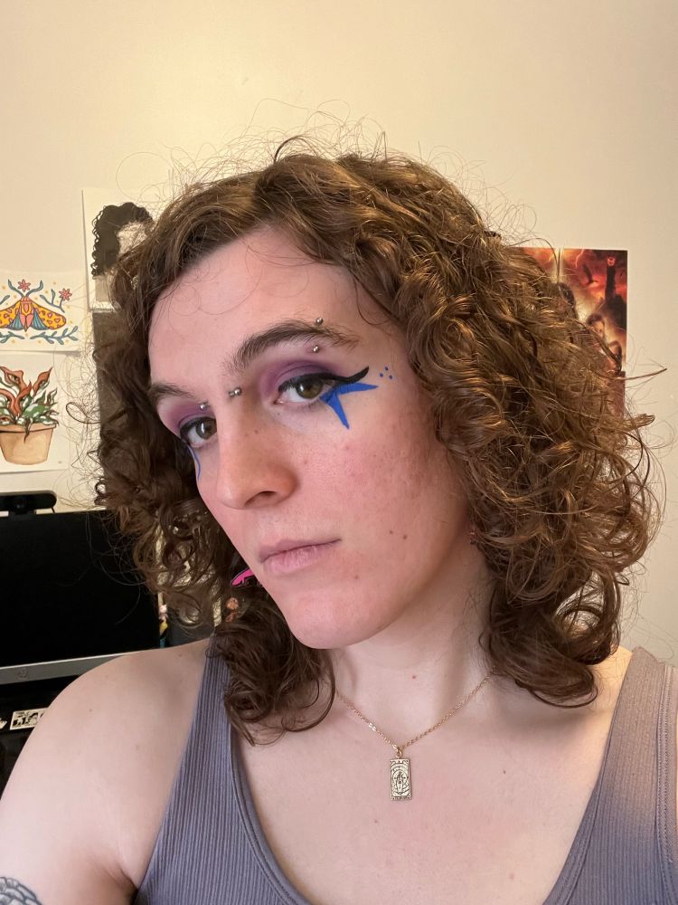 selfie of sadie, she has very curly hair, purple eyeshadow, black eyeliner, and two blue spikes painted on coming down from her eyes. also an eyebrow and bridge piercing