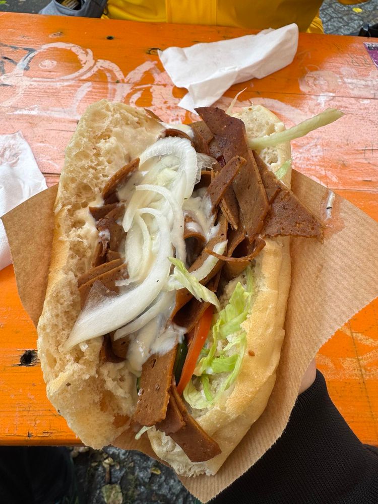 Ein veganer Seitandöner mit alles in naturbraunem Papier über einem orange bemaltem Holztisch, dahinter eine Papierserviette (war nötig, weil Stinkiki ging dann bis anne Ohren).