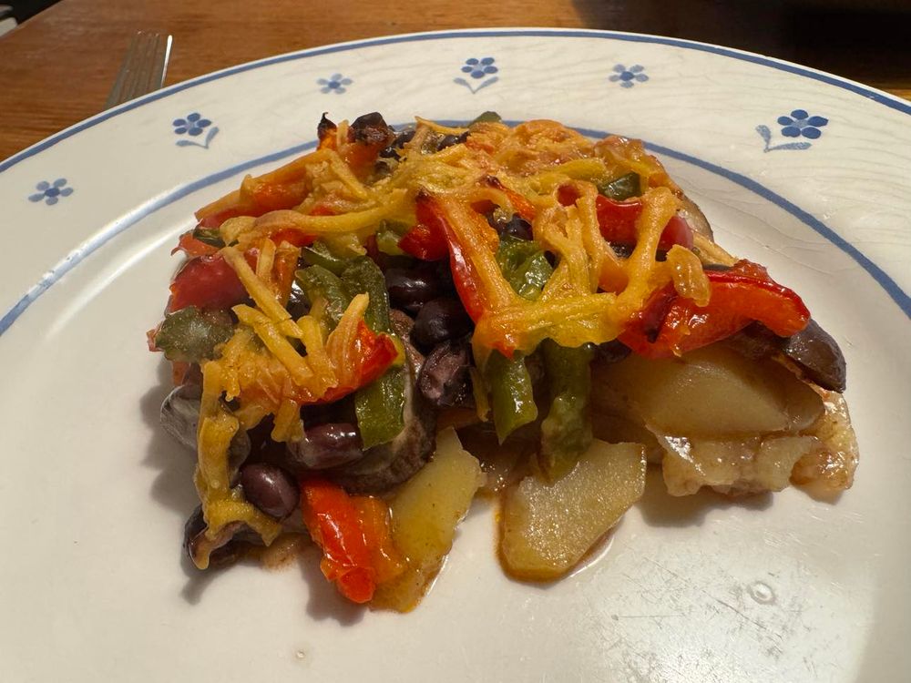 Der Auflauf aus dem anderen Bild auf einem flachen weißen Teller mit blauem Blümchendekorrand. Hier sind unter der Käseschicht Paprika, Kidneybohnen und Kartoffeln zu erkennen.