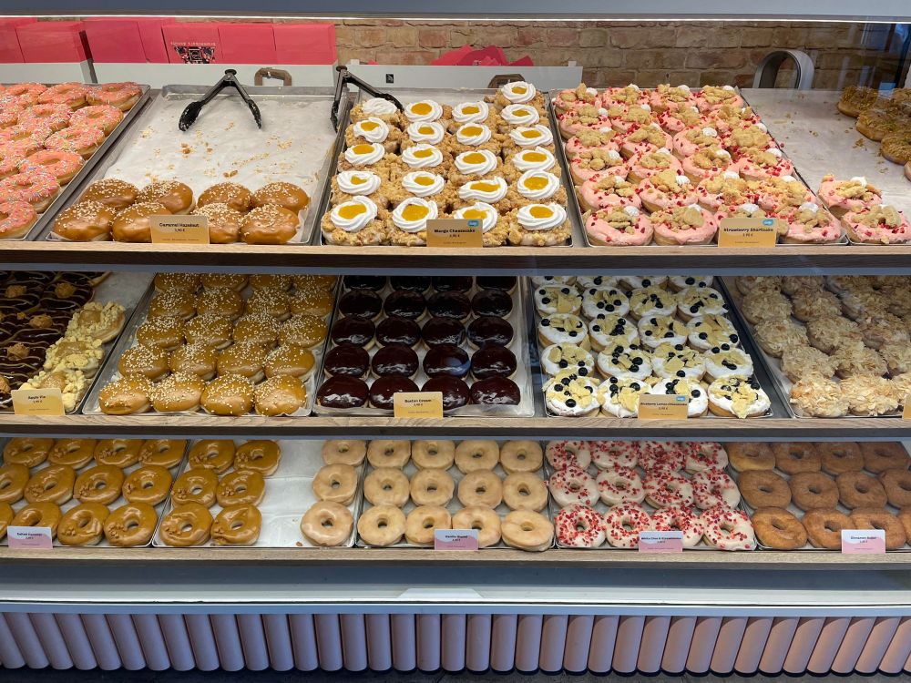 Eine Bäckereitheke mit vielen verschiedenen Donuts: gefüllt, ungefüllt, mit Streuseln, mit Cremegedöns, mit Fruchtgedöns, mit Zuckerguss, mit Mandeln, mit Zimtzucker, mit Schoki...