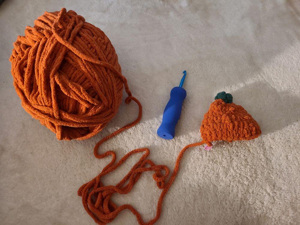 A skein of Bernat Blanket yarn, in the color Orange Leaf, against a white background. It's partially unwound, connected to a work in progress crochet project that resembles a pumpkin with a green stem. A blue 7.0 mm crochet hook with a blue foam grip rests between the two pieces.

Handmade by VexingHollow. Pattern (combination) by Cable and Canvas.