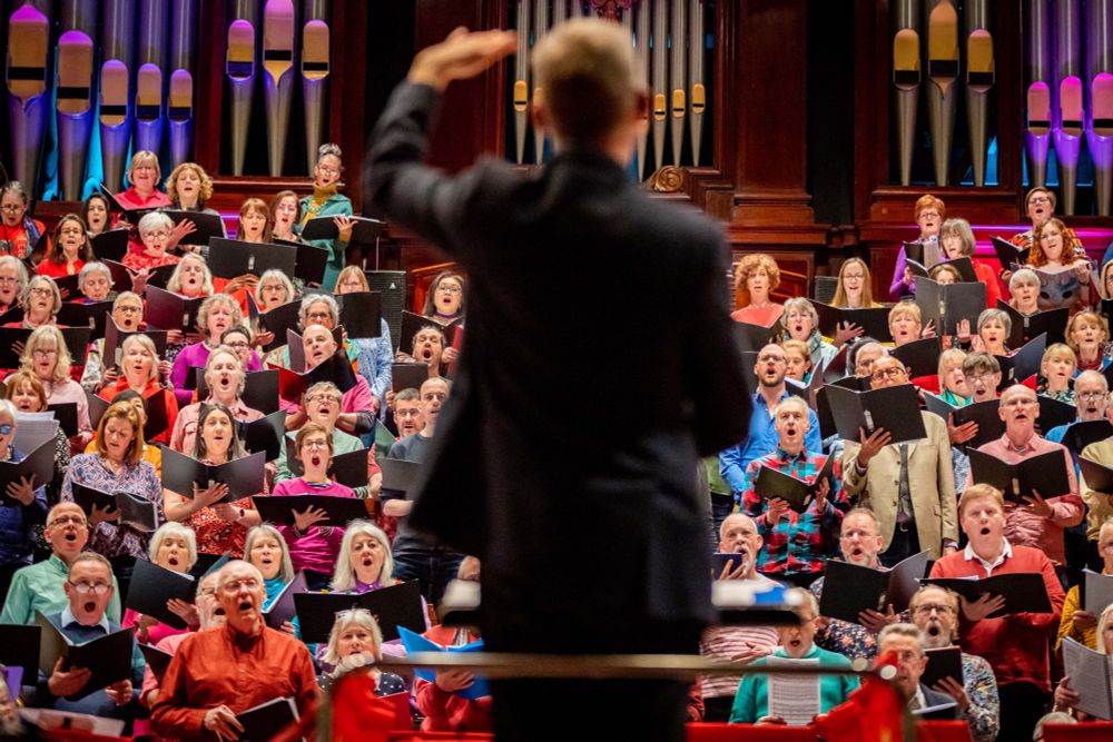 A large choir dressed in colourful attire performs under a pipe organ, led by a conductor gesturing confidently from the front.