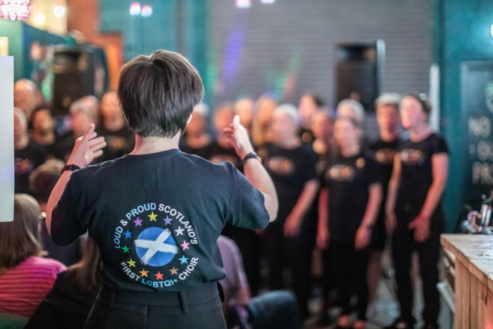 A conductor leads a choir wearing black shirts with a colourful logo, that reads 'Loud & Proud Scotland' celebrating Scotland's first LGBTQI+ choir.