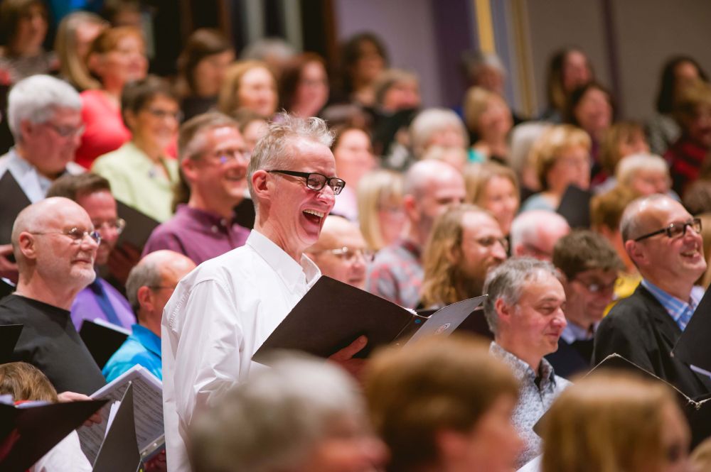 A diverse group of choir members stands on stage, holding music sheets, engaged happily in a performance in a well-lit auditorium.