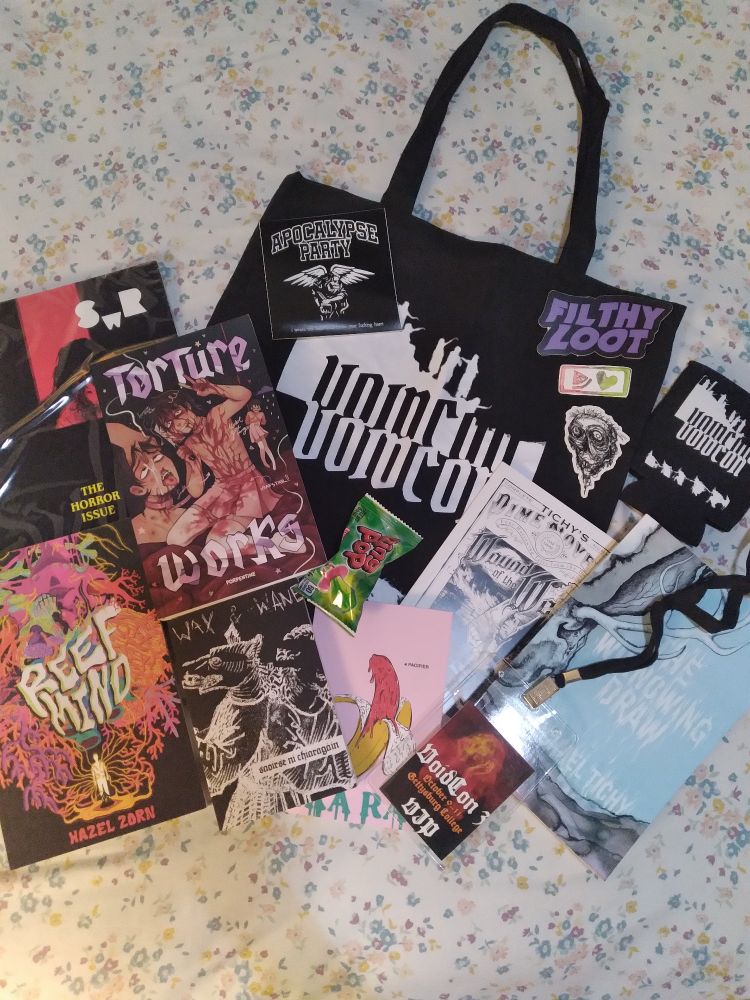 Photograph of an assortment of indie/punk horror books and merchandise arranged over a floral background. Books include Reef Mind by Hazel Zorn, Torture Works by Porpentine, Wax and Wane by Saoirse ni Chiaragain, Pacifier by Ira Rat, and The Winnowing Draw and Wound of the West by Michael Tichy. Merch includes the 2025 October horror issuse of The Southwest Review, a VoidCon tote bag, a VoidCon coozy, stickers from the publishers Apocalypse Party, Merigold, Filthy Loot and others, a VIP pass for the convention and a Ring Pop.