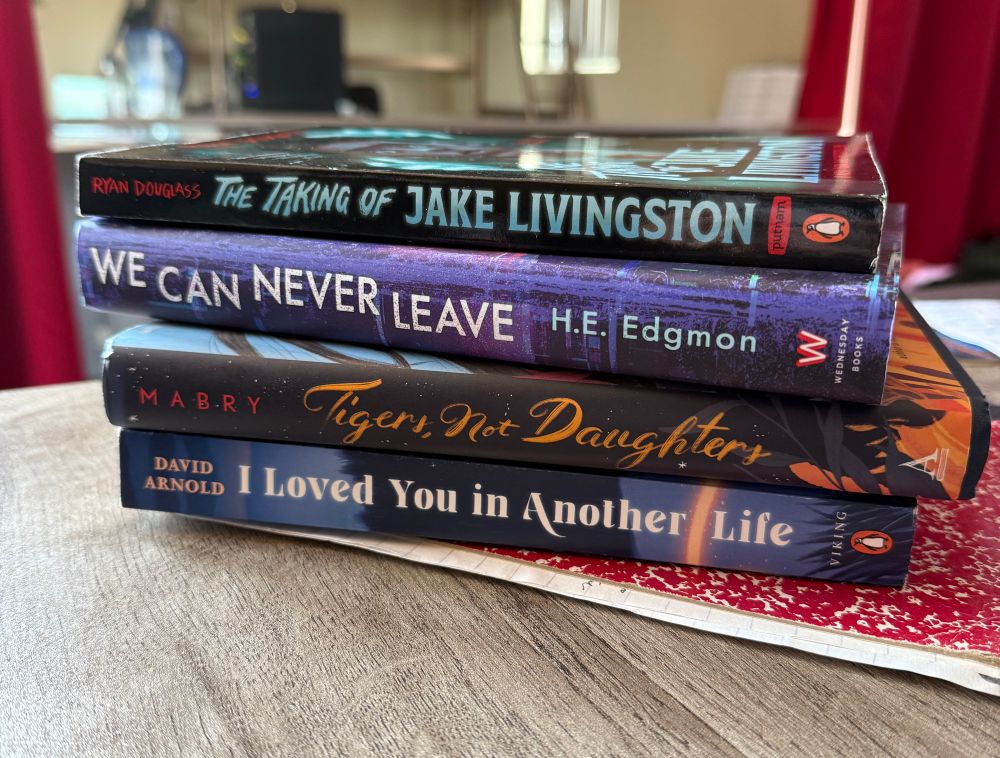 A stack of books. Includes The Taking of Jake Livingston by Ryan Douglass, We Can Never Leave by H.E. Edgmon, Tigers, Not Daughters by Samantha Mabry, and I Loved You in Another Life by David Arnold