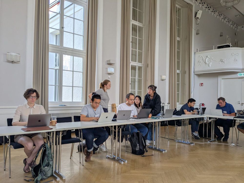 A group of people sitting at a long table in a bright room with large windows. Some are using laptops while others are standing.
