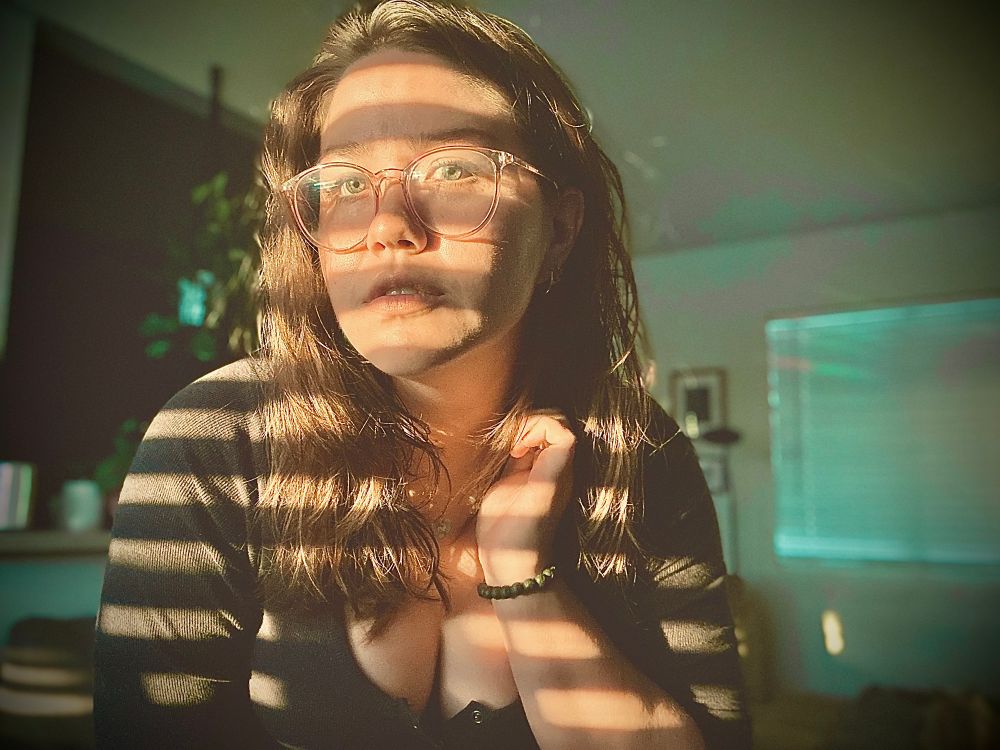 a self portrait photo of tay in her living room. the west facing window is letting stripes of light in, creating a pattern on her face. it highlights her green/hazel eyes