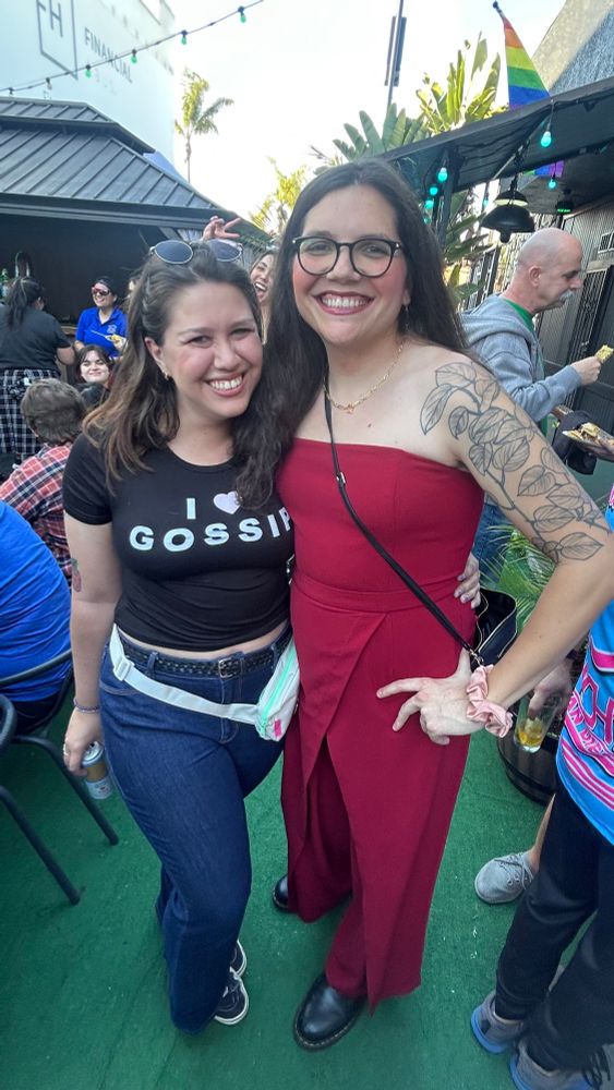 tay and ava at a gay softball event. ava looks amazing in a red strapless jumpsuit and tay has a black crop top that says "I 🩷 gossip" and blue jeans