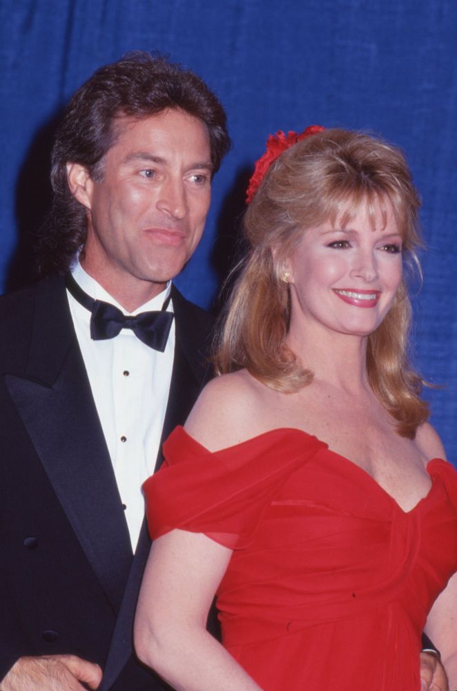 Professionally shot 1993 portrait of Deidre Hall and Drake Hogestyn at the 20th Annual Daytime Emmy Awards. Deidre wears a bright red off-the-shoulder dress with a soft ruffle neckline, her shoulder-length blonde hair styled in gentle layers and partly tied up with a matching red ribbon. She holds a gentle sideways smile, exuding elegance and warmth. Drake stands beside her in a classic black tuxedo with a white dress shirt and black bow tie, his hair slightly longer at the back in true early-90s style. Both are looking slightly away from the camera, framed by a rich blue curtain backdrop. The emotional tone is nostalgic and glamorous, celebrating their legacy as Marlena Evans and John Black from Days of Our Lives, especially during the height of their iconic affair storyline in 1993.
