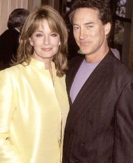 A candid early 2000s photo of Deidre Hall and Drake Hogestyn, best known for their roles as Marlena Evans and John Black on Days of Our Lives. Deidre wears a pale yellow silk skirt suit with a light-colored top underneath, and her shoulder-length light brown hair is softly layered with bangs. Drake stands beside her, wearing a dark pinstriped suit jacket over a lilac t-shirt. Both are smiling gently and looking directly at the camera. The background suggests a softly lit indoor event with other attendees. The image captures warmth, nostalgia, and the enduring glamour of two beloved soap opera icons.
