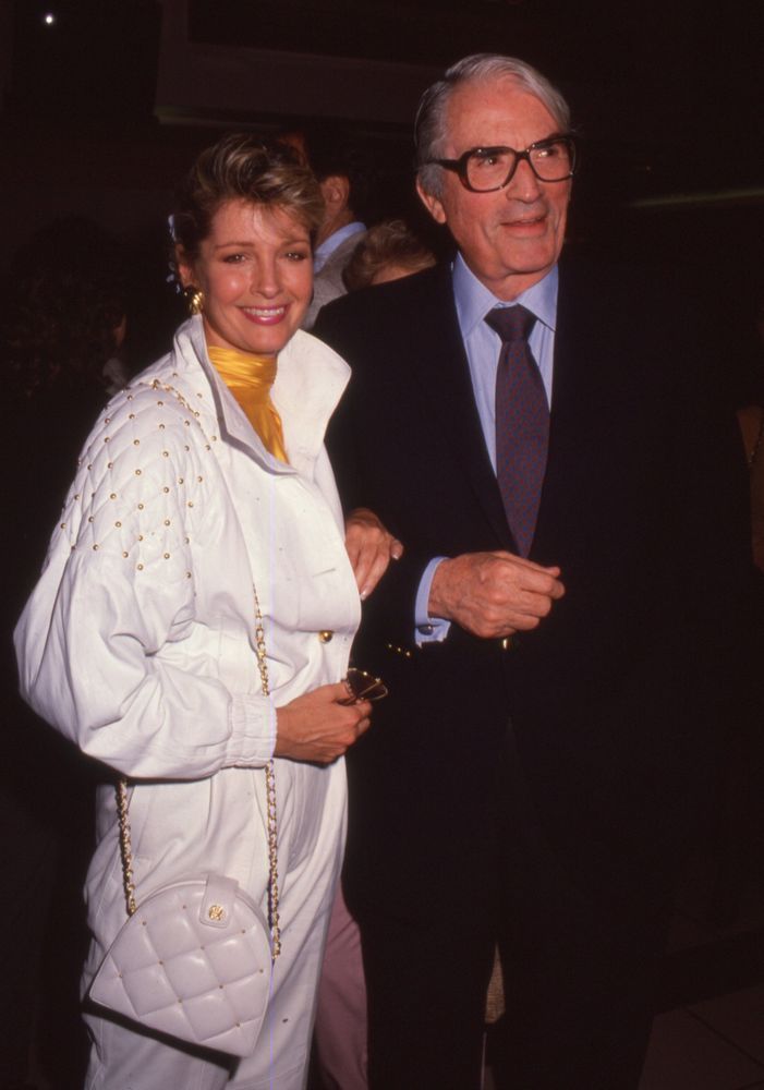 Late 1980s photograph of Deidre Hall and a man believed to be Gregory Peck at a formal event. Deidre wears a white jumpsuit with gold embellishments on the sleeves, a yellow scarf tied around her neck, and carries a white quilted handbag with a gold chain strap. Her softly layered, shoulder-length blonde hair is styled with volume and elegance. She smiles warmly, standing beside a man in a dark suit, light blue shirt, and patterned tie. The background is dimly lit, suggesting an indoor gala or premiere. The emotional tone is nostalgic and glamorous, capturing Deidre’s transition from daytime icon as Marlena Evans on Days of Our Lives to her nighttime guest-starring era in shows like Wiseguy, Murder, She Wrote, and Columbo. The image reflects legacy, poise, and the quiet thrill of Hollywood-meets-daytime royalty.
