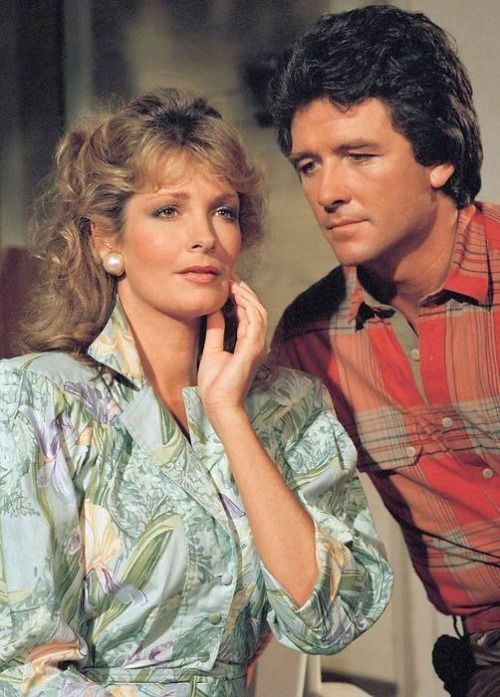 1986/87 promotional or behind-the-scenes image from Our House, featuring Deidre Hall and Patrick Duffy. Deidre wears a light blue floral-print dress with prominent shoulder pads and large pearl earrings. Her blonde hair is curly and partly pinned up, styled in a voluminous mid-80s fashion. Patrick stands beside her in a red and brown plaid shirt layered over a grey undershirt. Deidre gazes into the distance with a contemplative expression, while Patrick looks directly at her with quiet intensity. The background is softly blurred, suggesting an indoor set. The emotional tone is tender and reflective, capturing their roles as Jessie Witherspoon and her late husband. Legacy context highlights Deidre Hall’s fame as Marlena Evans on Days of Our Lives and Patrick Duffy’s iconic role as Bobby Ewing on Dallas.
