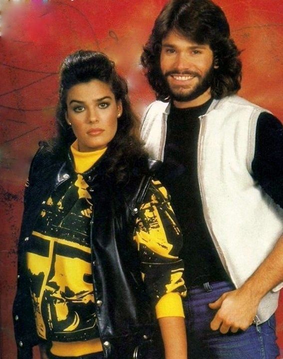 Kristian Alfonso and Peter Reckell in a vibrant early 1980s promotional portrait, likely from 1983. Alfonso wears a black leather jacket over a bold yellow-and-black graphic sweatshirt, while Reckell sports a white vest layered over a black shirt and purple trousers. Set against a vivid red backdrop with abstract patterns, the duo exudes youthful charisma and the magnetic chemistry that would define their iconic pairing on Days of Our Lives.
