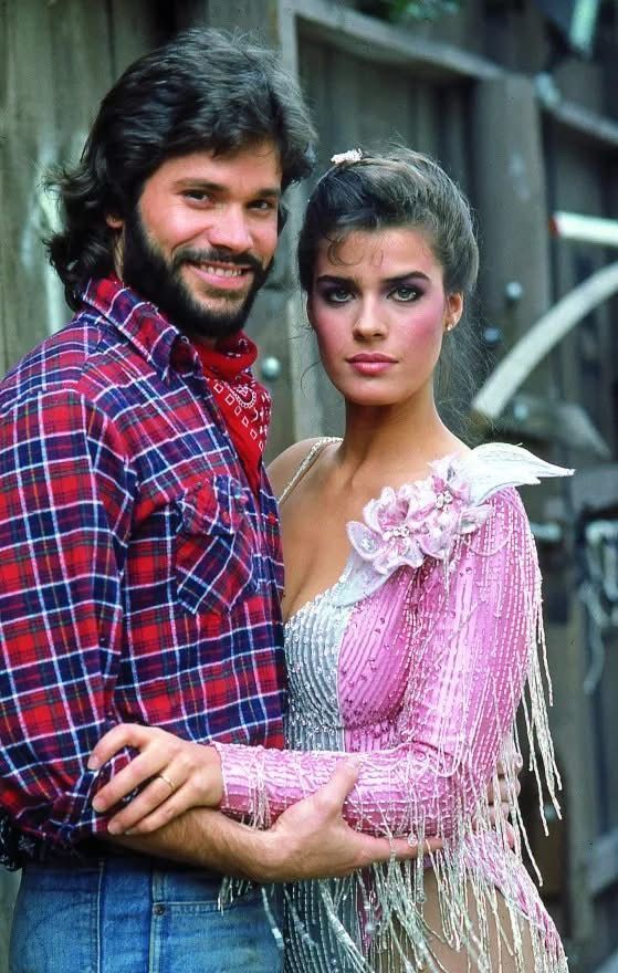 Kristian Alfonso and Peter Reckell in a playful early 1980s outdoor portrait. Alfonso wears a pink-and-white sequined Western-style outfit with fringe and floral embellishments, while Reckell stands beside her in a red plaid shirt and red bandana. With his arm around her waist and her hand resting on his, the duo pose in front of a rustic wooden fence, radiating charm and the spirited chemistry that made Bo and Hope a beloved soap opera couple.
