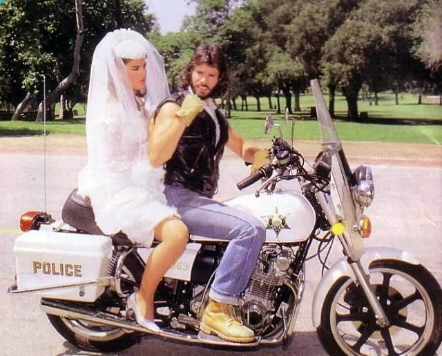 Kristian Alfonso and Peter Reckell in a dramatic 1984 scene from Days of Our Lives. Alfonso, in a white wedding gown and veil, rides pillion on a police motorcycle behind Reckell, who wears a sleeveless leather vest, jeans, and boots. Set in a leafy outdoor location, the image captures the moment Bo dramatically interrupts Hope’s wedding to Larry Welch—an iconic twist in their tumultuous, legendary love story.
