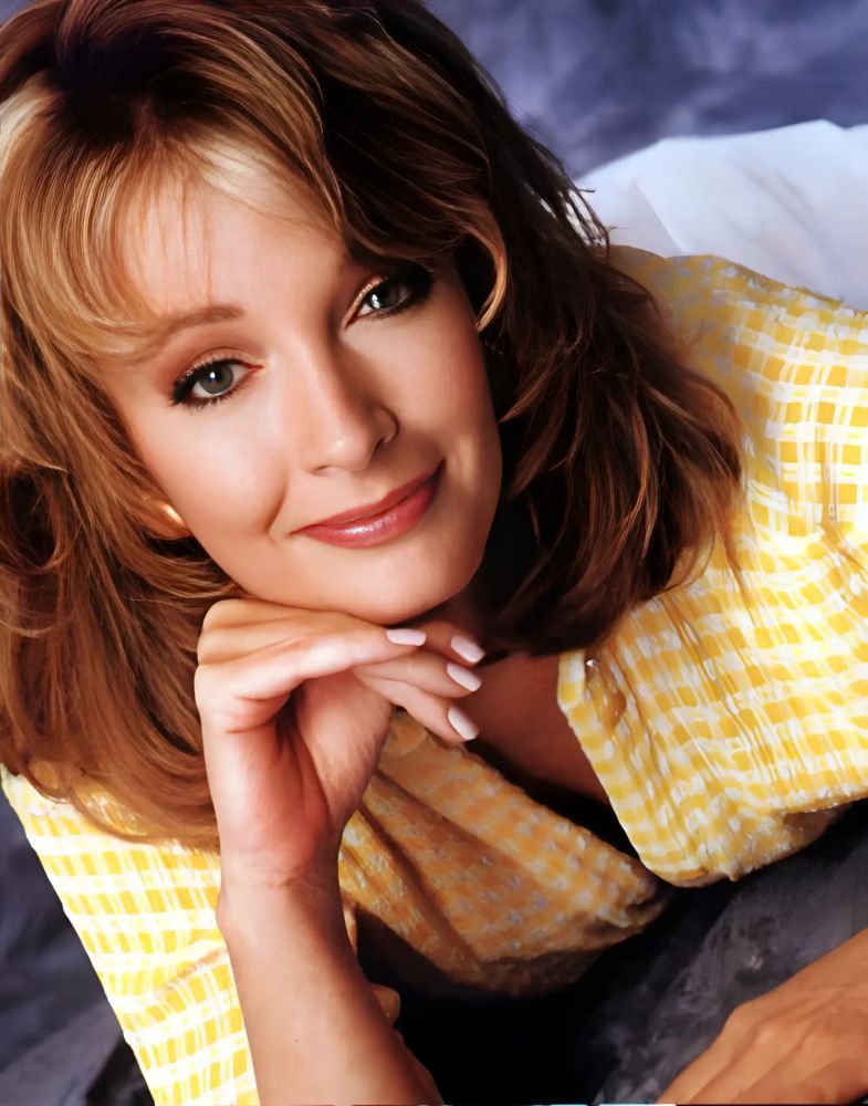 A professionally shot 1994 portrait of Deidre Hall, best known for her role as Marlena Evans on Days of Our Lives. She wears a white and yellow checkered top, possibly the same one featured in the Smith Island scenes with John Black during the possession storyline. Her shoulder-length light brown hair is softly layered and styled in gentle waves. She rests her chin on her fingers and looks directly at the camera with a serene, knowing smile. The background is softly blurred in shades of blue, creating a calm and intimate atmosphere. The image captures a moment of poised elegance and emotional depth, just before her character’s transformation into the iconic MarDevil.
