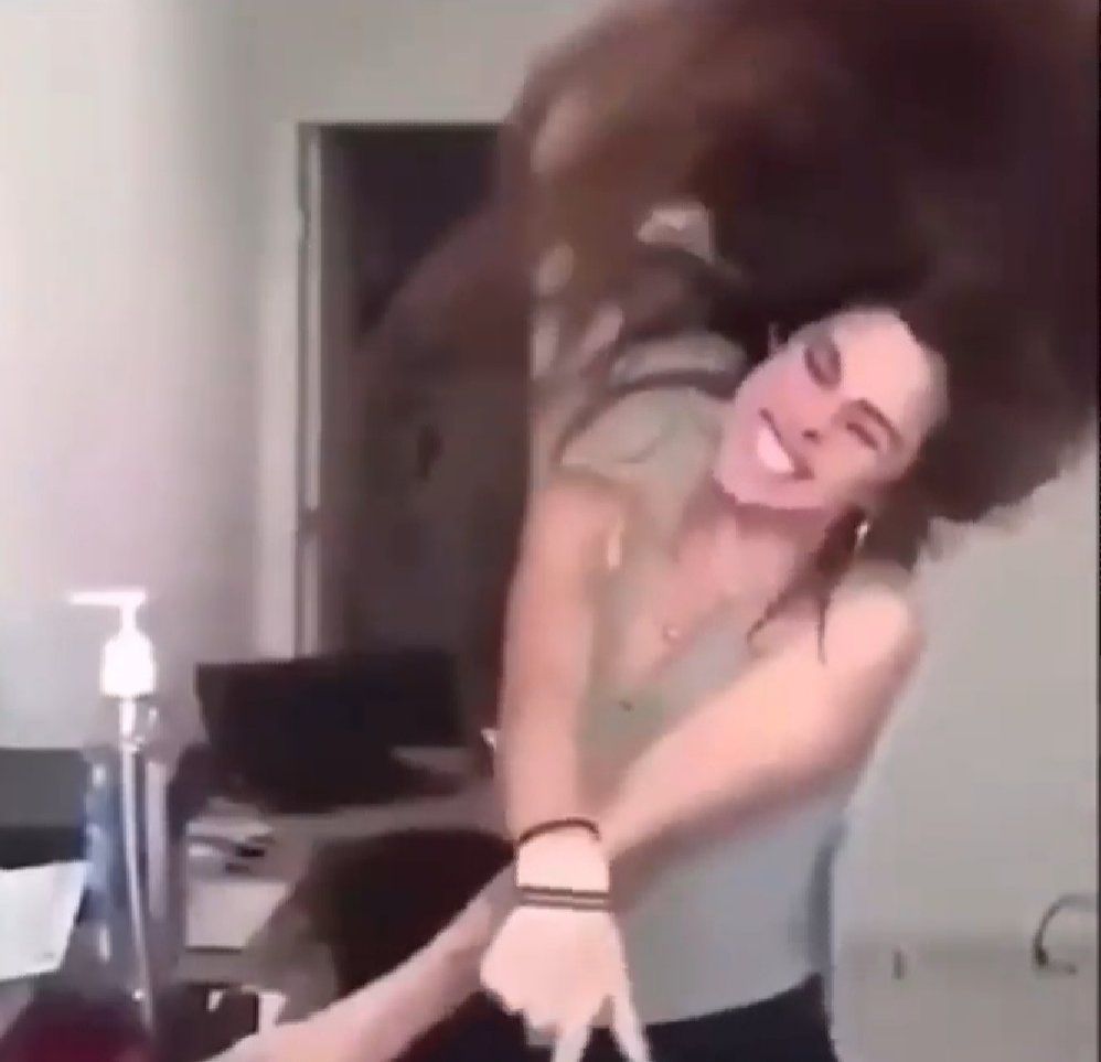 Mulher vestindo calça preta e blusa acinzentada, sorrindo e jogando o cabelo