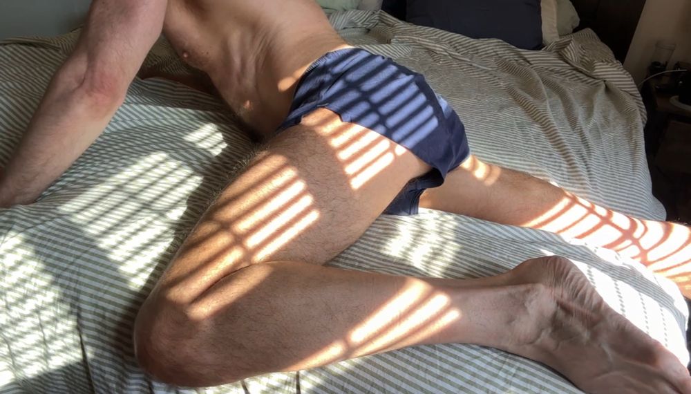On a bed in short shorts with light coming through slats in blinds