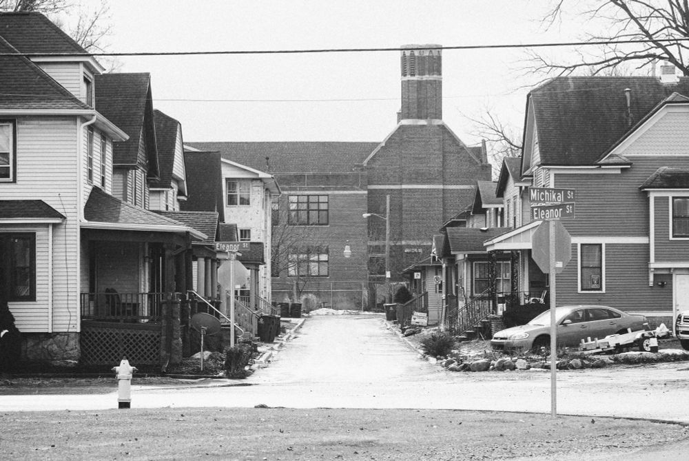 This photo captures a quiet street scene in the Stuart Area Historic District of Kalamazoo, Michigan. The view shows two rows of early 20th-century houses with porches and well-maintained yards, all framed by the bare trees of early winter. At the end of the street, the large, red-brick St. Augustine Cathedral Church is visible, standing tall behind the row of houses. The photo is in black and white, adding a timeless, peaceful feel to the scene. The street signs for "Michikal St" and "Eleanor St" are prominent in the foreground, with a quiet, snow-covered street extending toward the church.