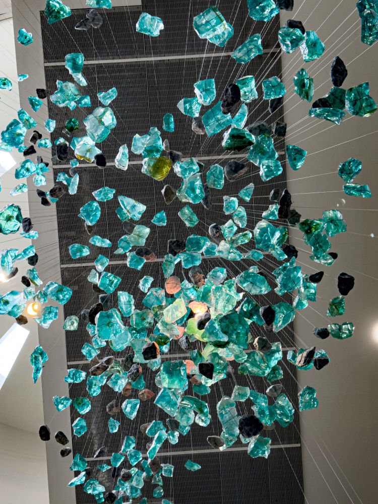 Looking upward at a large suspended sculpture composed of dozens of translucent aqua-blue and green resin shapes, interspersed with darker forms, all hanging on fine wires from the ceiling. The pieces appear to float like a frozen cascade of water or a river in midair. Bright natural light and spotlights illuminate parts of the installation, creating reflections and shadows against the tall architectural ceiling.
