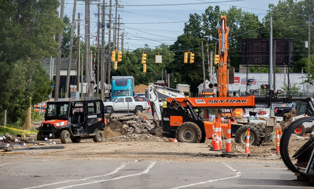 Road construction scene on North Westnedge Avenue in Kalamazoo, Michigan. The foreground shows broken pavement and dirt, with construction equipment including a black-and-orange utility vehicle and a large orange SkyTrak telehandler. Traffic cones are scattered throughout the site. In the background, traffic lights hang over the busy intersection, and several vehicles wait at a railroad crossing, including a teal semi-truck and a white pickup. Tall utility poles and overhead wires fill the sky, and trees and commercial buildings frame the scene.