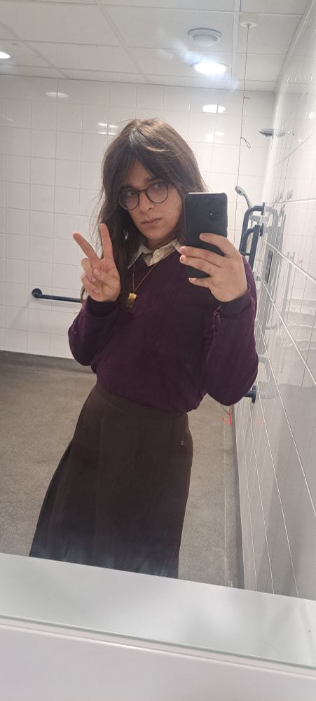 a nonbinary person taking a selfie in a bathroom  mirror. they are wearing a brown skirt and a purple jumper with the collar of their shirt peeping out over it. they are making a peace sign