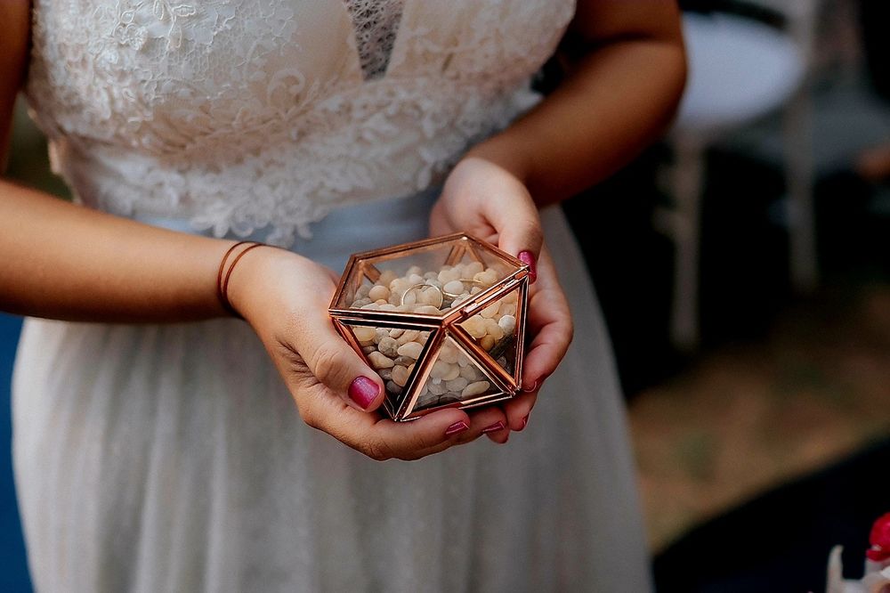 Noiva vestida de branco, com unhas cor fúcsia, segurando um porta alianças de vidro, cheio de pedras brancas, com duas alianças douradas em cima
