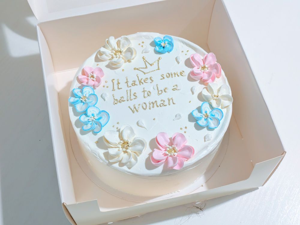 White round cake with pink, white and light blue flowers and the text that says "it takes some balls to be a woman".
