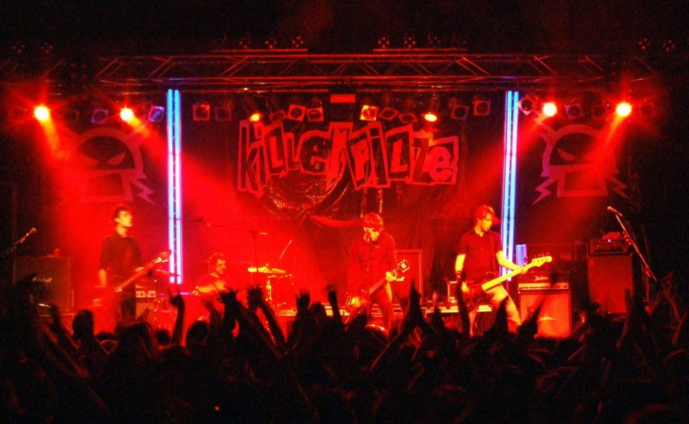 Bühnenfoto eines Auftrittes der Killerpilze im LKA Stuttgart. Im Vordergrund dunkel viele hochgereckte Hände, die Bühne in tiefem Rot. Mittig oben das Killerpilze Logo. Man kann die vier Musiker erkennen, aber das Bild ist insgesamt nicht sehr scharf, deshalb fehlen Details. 