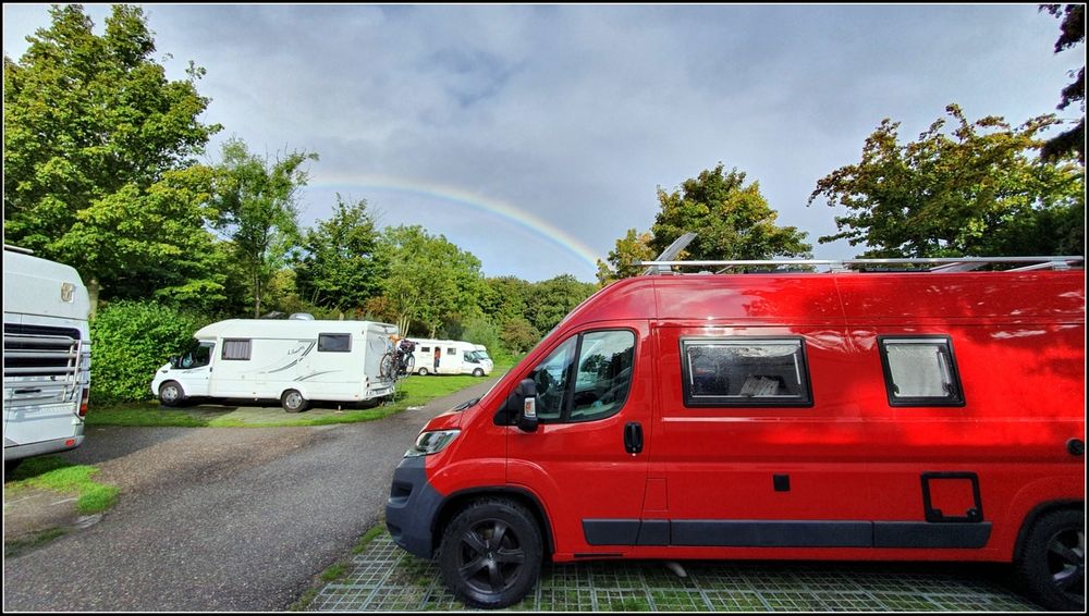 Ein Campingplatz mit mehreren Wohnmobilen auf Rasensteinen. Rechts im Bild steht ein rotes Kastenwagen-Wohnmobil. Im bewölkten Himmel ist ein Regenbogen zu sehen. 