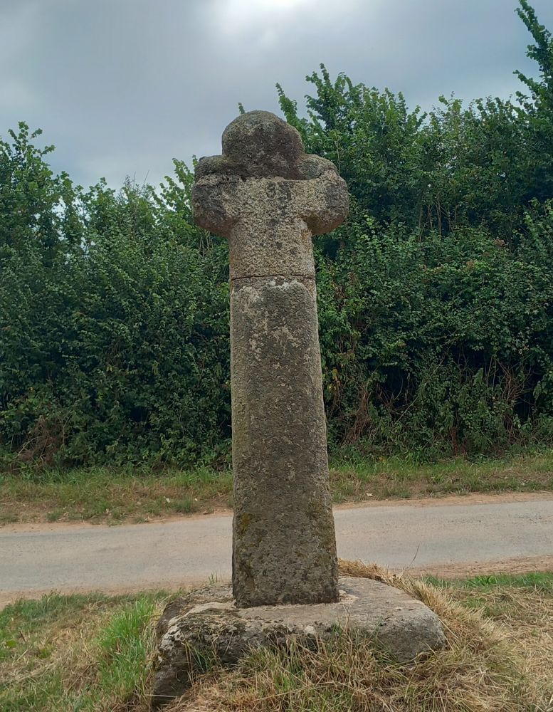 A photograph of an old stone cross, with an unusual rounded head, by the side of a country lane.