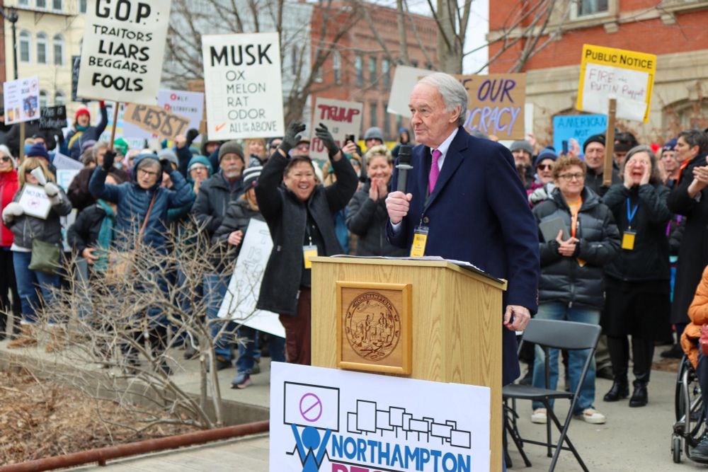 Senator Markey speaks behind a podium with a crowd behind him carrying signs.