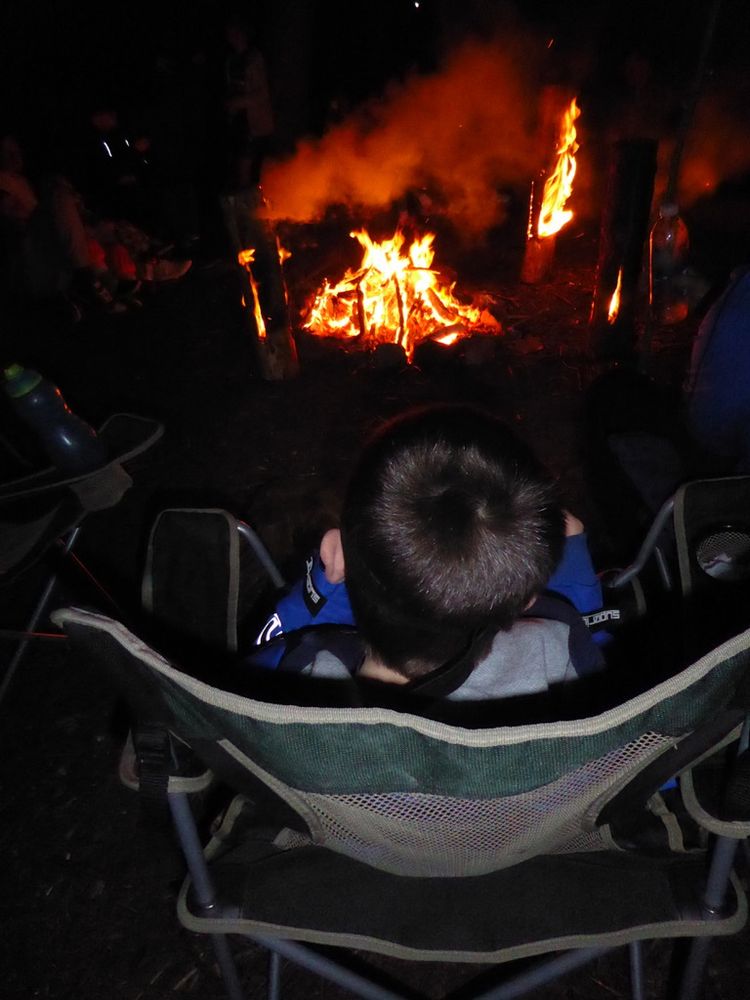 This is an image of my young son on his first camping trip.  The photo is shot from behind him, looking over his head as he sits in a camping chair staring intently at the blazing camp fire and upright fire logs.