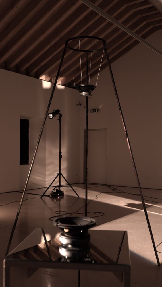 View of the installation “Immanent Field (VVV)”. In the photo we can see a loudspeaker set on a plinth in the foreground, behind it a two-and-a-half-metre high piramidal metallic structure from where another loudspeaker hangs and, in the background, a three-metre tripod with a spotlight.