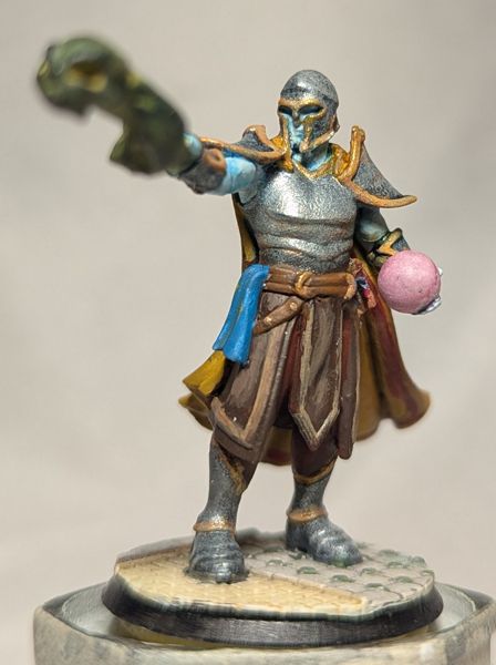 A painted miniature depicting a wizard casting a necromantic spell ending in a skull, emanating from an outstretched right hand.

His ghostly skin is a pale blue/white, and a pinkish crystal ball is cradled in his left hand.  He wears a helmet, breastplate, pauldrons, greaves, and armored boots of silver with gold trim.

A brown leather skirt and tabard hang from his waist, over red-brown trousers.  Hanging from his leather belts is a blue cloth patterned in yellow stars on his right hip, and a spell book upon the left.  Draped from his shoulders is a yellow cloak with a red border around the interior face.

The image is a frontal view.