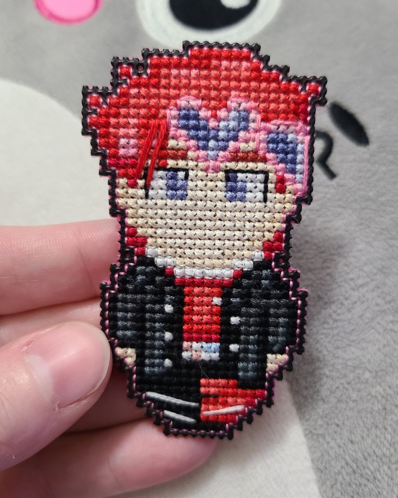 a small cross stitch character with red hair, pink heart goggles, and black and red clothes