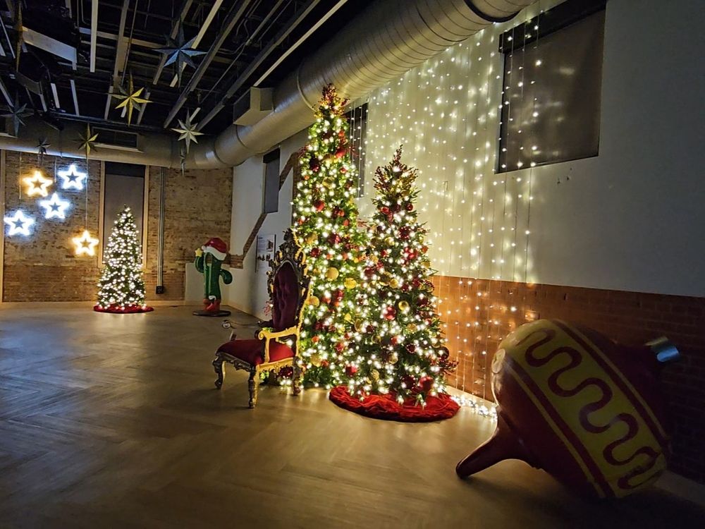 The same large, dark, and empty room. Three Christmas trees with lights. There is a very large Christmas ornament as a decoration on the ground next to two of the trees. There is a fancy gold and velvet-like chair in front of the trees.