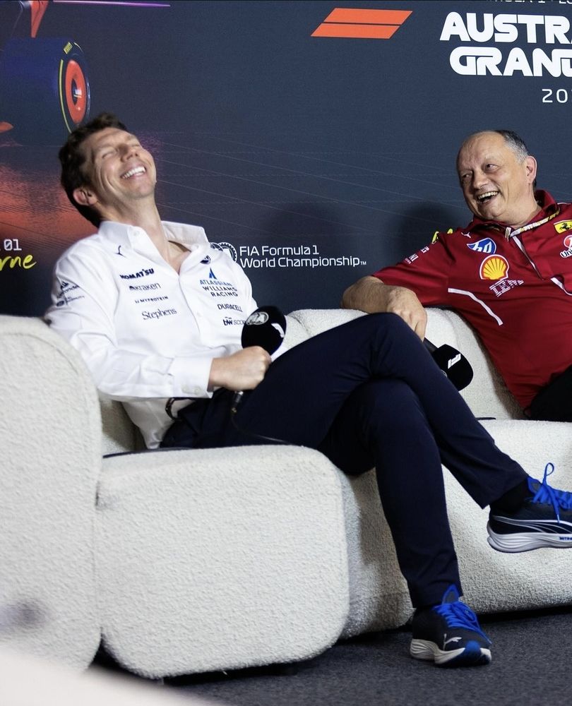 A photo from James Vowles' insta jv.f1. He wears a white shirt with the Williams logo and sponsors, dark jeans and black trainers with bright blue laces. Fred Vasseur is next to him, wearing his red Ferrari top, and looking over at him with a laugh. James' head is thrown back in gleeful laughter and his eyes are closed, two open buttons exposing his collarbone and long neck. 