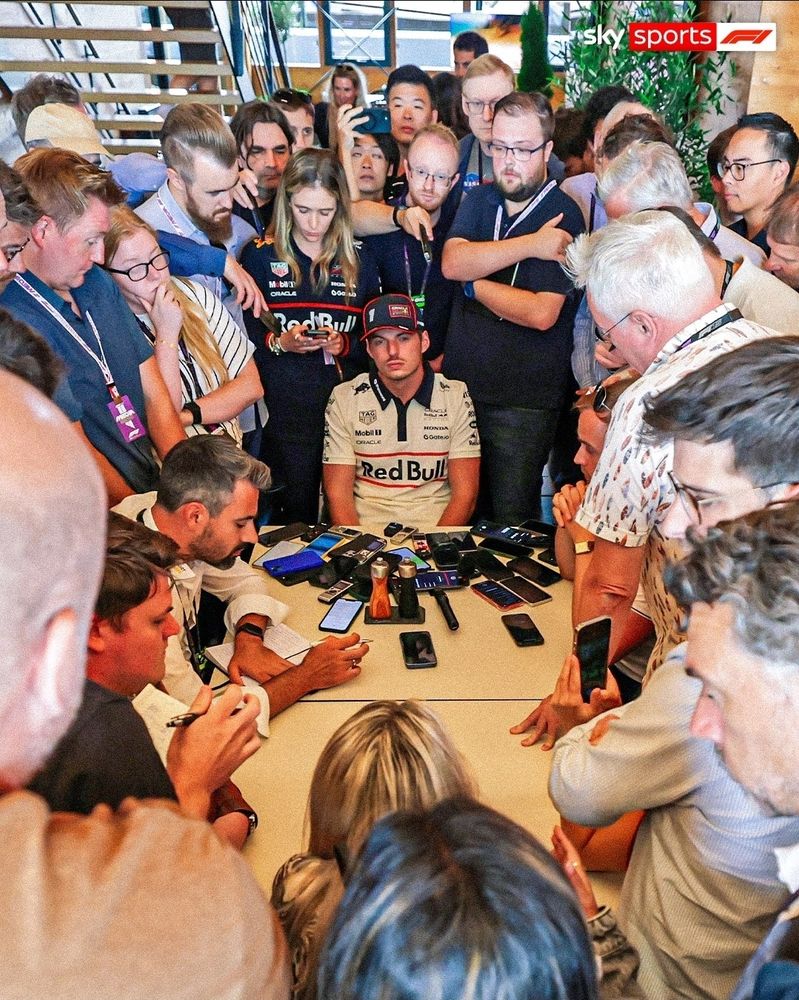 From skysportsf1 on Instagram.  Max Verstappen wears a white Red Bull team shirt and blue Red Bull cap. He is absolutely surrounded by journalists with a pile of mobile phones and recording devices in front of him. He has a sour, murderous expression on his face. 