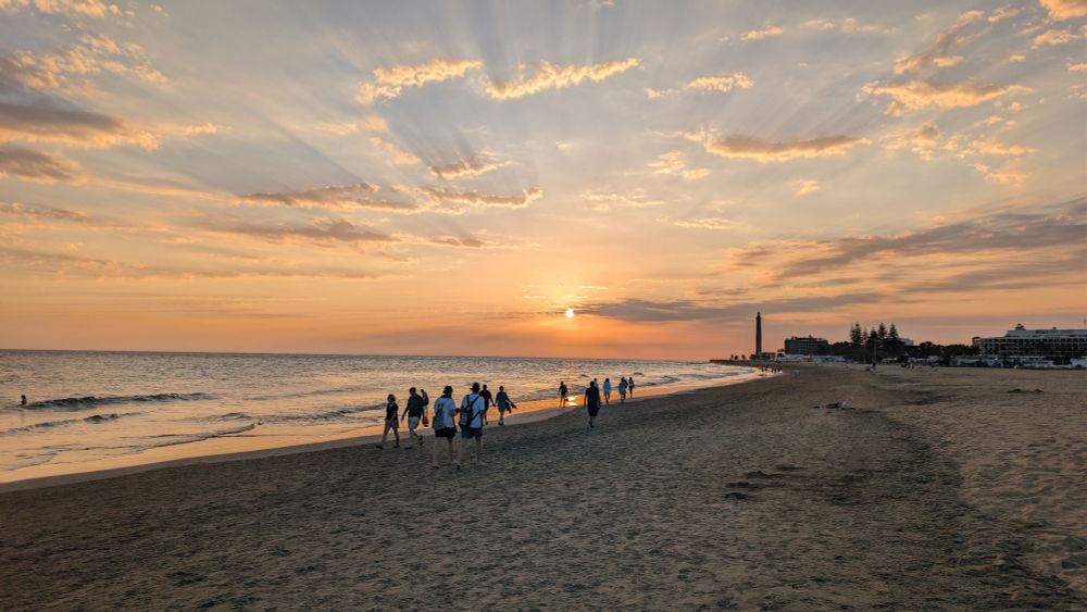 People walking along the beach. In the distance the lighthouse stands out. The orange sun is low enough that the clouds are casting shadows across the sky like sun beams. 