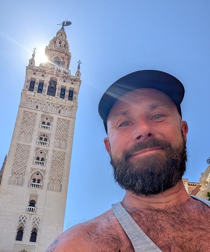 Me smiling into the camera. Wearing a cap and a vest. Lots of chest hair on show. Seville cathedral tower behind me. The sun is behind the tower. The sky is pure blue.