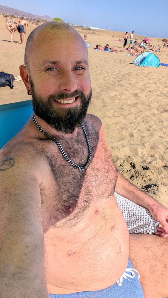 I'm wearing a steel necklace and blue budgie smugglers. Sitting on a deckchair with the beach and blue sky behind me. Several surgery ports on my belly are healing nicely. 