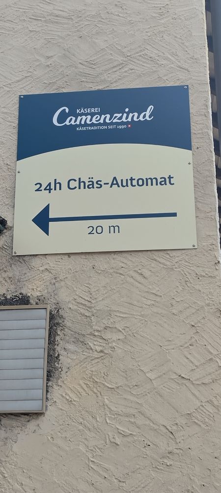 Ein Schild auf dem steht oben Käserei Camenzind Kösetradition seit 1990 Darunter steht 24h Chäs-Automat (24h Käse-Automat) <------------------ 20m